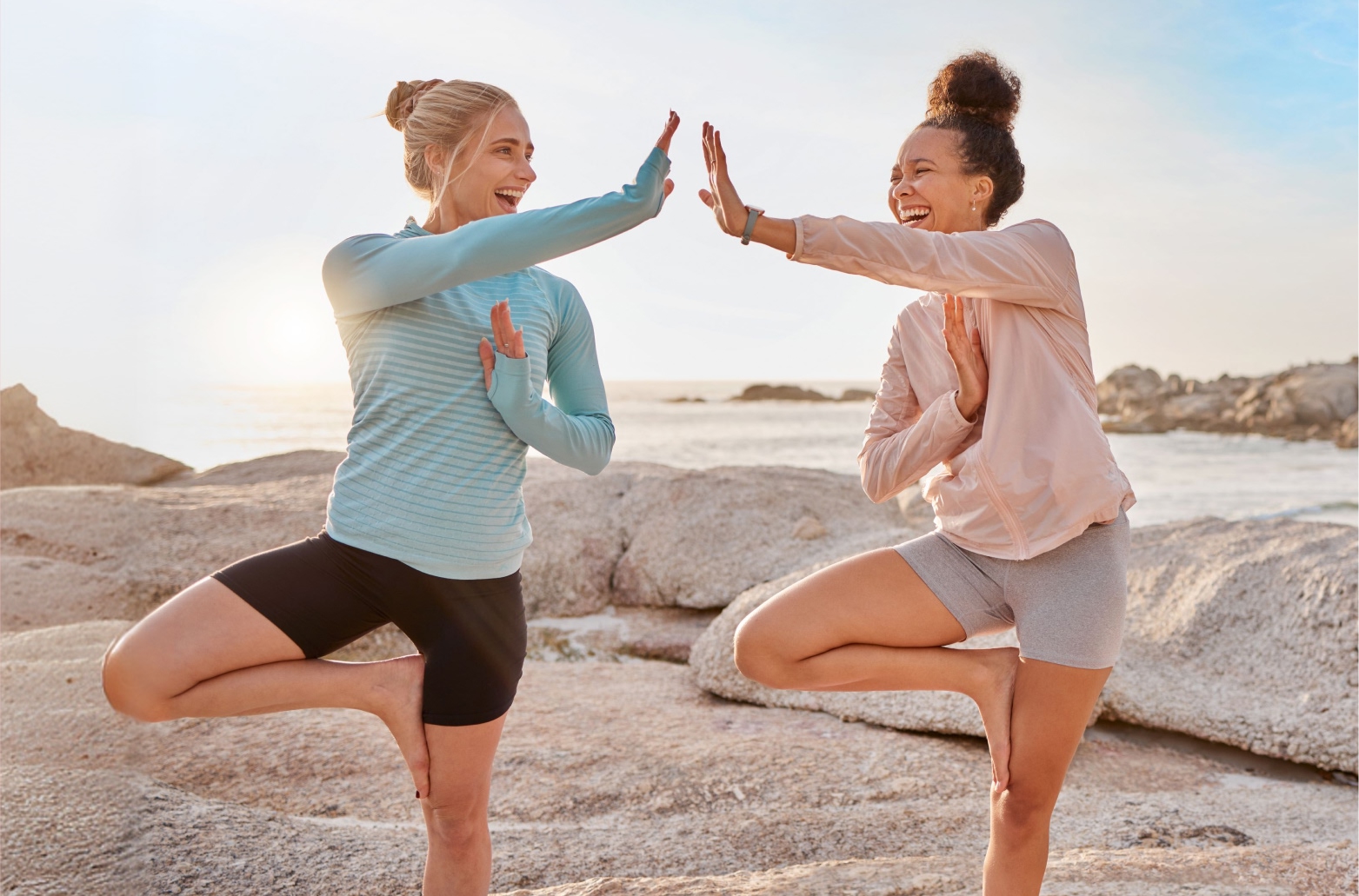 Две улыбающиеся девушки выполняют позу дерева (Врикшасана) на скалах у моря — иллюстрация к разделу «Частые ошибки новичков в йоге и пилатесе»